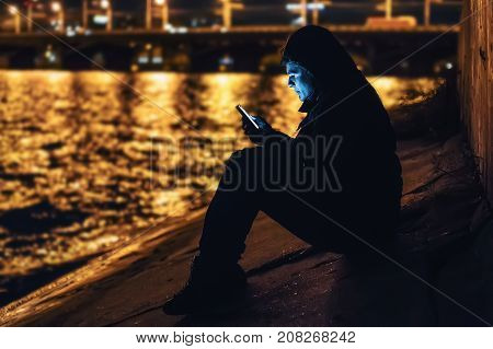 Sad lonely man sitting on pier with smartphone in hand at blurred sea background with reflection of city night lights, depression and loneliness concept, dark toned