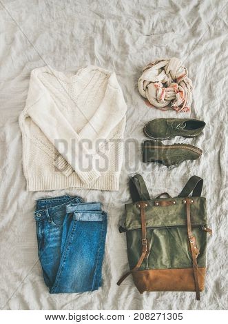 Flat-lay of Fall lady' s clothes outfit. Ecru colored knitted woolen sweater, linen scarf, denim jeans, haki suede boots, backpack over grey linen bed blanket, top view, copy space. Trendy Autumn look