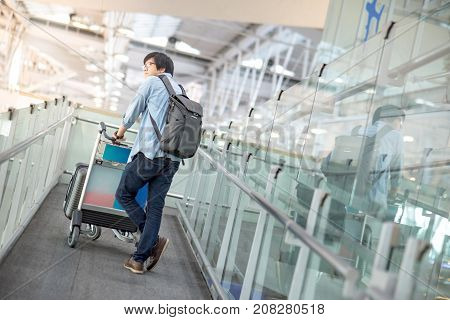 Young Asian man walking with airport trolley and his suitcase luggage in the international airport terminal arrival from travel abroad