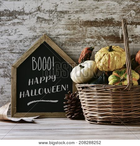 a house-shaped chalkboard with the text booo happy halloween, and a worn wicker basket with an assortment of different pumpkins on a rustic wooden table