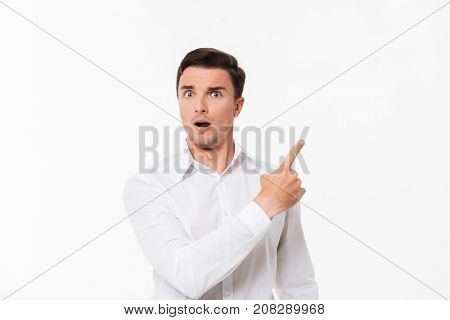 Close up portrait of a surprised astonished man in a white shirt standing and pointing finger away at copy space while looking at camera isolated over white background