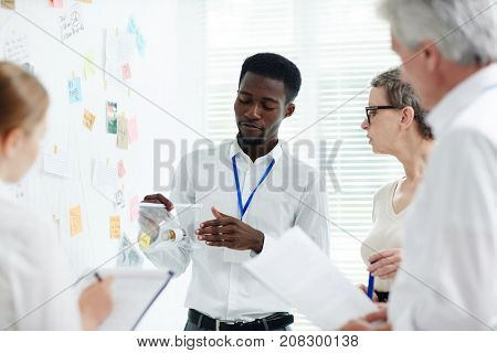 Handsome African American police officer showing evidence taken from crime scene to his team while having discussion at modern open plan office