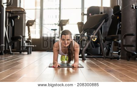 Front view of a young fit woman smiling while practicing the forearm plank exercise for core strength indoors at the gym