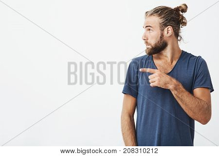 Close up portrait of joyful handsome guy with trendy haircut and beard in profile pointing his finger at white background with copy space for advertisement