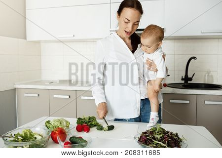 Beautiful young mother holding little son on hands in kitchen showing him how to cut vegetables for salad. Child looking at mother hands with big interest