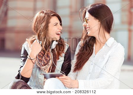 Two young and beautiful girl sitting at the street and watching something on a tablet.