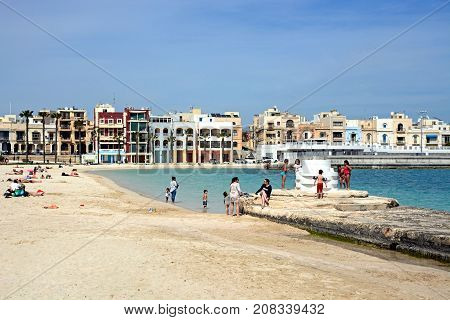 BIRZEBBUGA, MALTA - APRIL 1, 2017 - Tourists relaxing on the beach with waterfront buildings to the rear Birzebbuga Malta Europe, April 1, 2017.