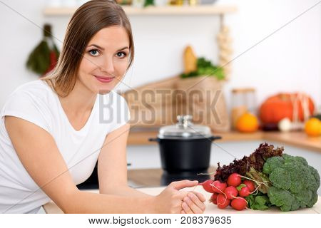 Young cheerful smiling woman is ready for cooking in a kitchen. Housewife sitting at the table and looking at the camera.