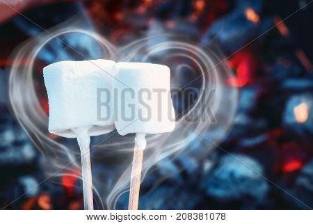 Two white sweet marshmallows roasting over fire flames. Smoke in form of heart. Marshmallow on skewers roasted on charcoals. Sweet love concept