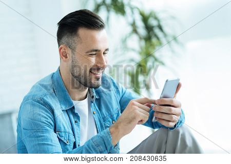 Innovative generation. Side view on a happy millennial guy sitting indoors and smiling cheerfully while touching a screen of her phone alone.