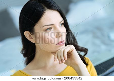 Focusing on dreams. Side view on a serious young woman looking into vacancy and resting her chin on a hand while dreaming about something.