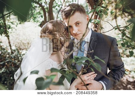 Beautiful Gorgeous Bride And Groom Kissing In Sunny Spring Park Among Green Leaves And Blooming. Hap