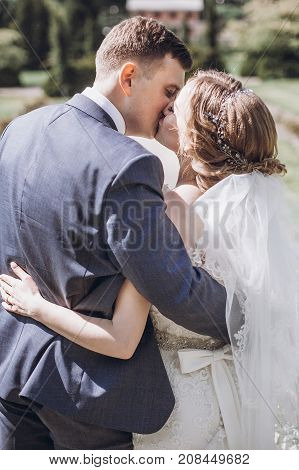 Beautiful Gorgeous Bride And Groom Kissing In Sunny Spring Park And Enjoying Day. Happy Wedding Coup