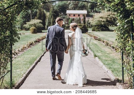 Beautiful Gorgeous Bride And Groom Posing In Sunny Spring Park Among Green Leaves. Happy Wedding Cou