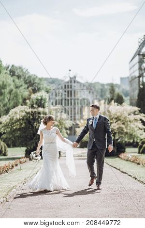 Beautiful Gorgeous Bride And Groom Posing In Sunny Spring Park Among Green Leaves. Happy Wedding Cou