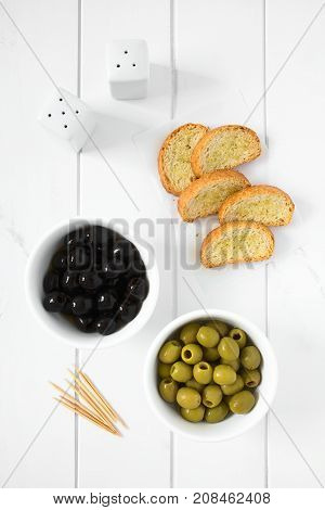 Pitted black and green olives in bowls with crostini on the side photographed overhead on white wood with natural light