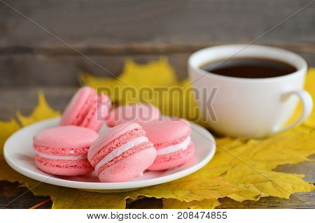 Delicious French macarons. Light pink macarons on a white plate, a cup of coffee, yellow leaves on an old wooden background. Fall breakfast concept. Closeup