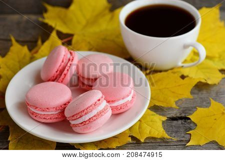 Sweet French macaroons. Light pink macaroons on a white plate, a cup of coffee, yellow leaves on an old wooden background. Autumnal breakfast concept. Closeup