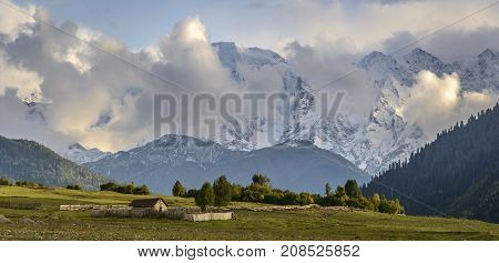 Little stone house surrounded with wooden fence on alpine meadow. High mountains covered with snow and clouds in background.