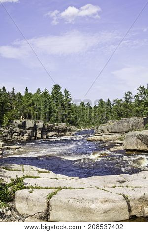 Vertical of rapids running between a rocky shoreline with a rock face in the foreground and surrounded by trees and greenery at Pabineau Falls near Bathurst, New Brunswick on a bright sunny day with blue skies and clouds in August.