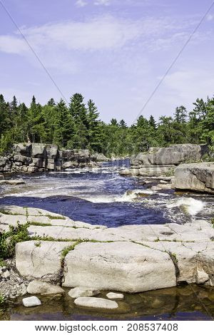Vertical of rapids running between a rocky shoreline with a rock face in the foreground and surrounded by trees and greenery at Pabineau Falls near Bathurst, New Brunswick on a bright sunny day with blue skies and clouds in August.