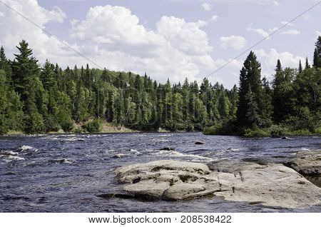 Wide view of a some small river rapids off a rocky shoreline surrounded by trees and greenery at Pabineau Falls near Bathurst, New Brunswick on a bright sunny day with blue skies and clouds in August.