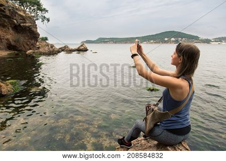Female tourist takes photo with cell phone sitting on the rock into Mediterranean Sea.