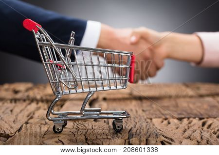 Close-up Of Shopping Cart On Wooden Desk In Front Of Businesspeople Shaking Hands