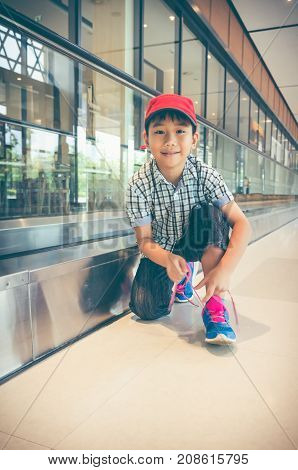 Happy Asian Child Sitting And Tying To Tie Shoelaces Near Walkway In Modern Airport.