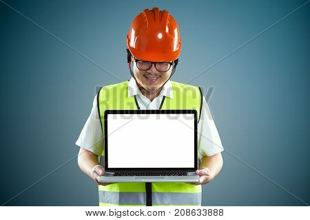 Young asian construction workers in safety helmet and using laptop with empty screen to show client something isolated on blue background .