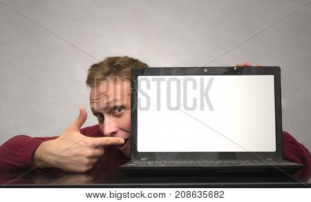 A smiling young man hiding behind laptop and showing by finger on blank screen of laptop computer on the table. Special offer. Big sale. Contact us.