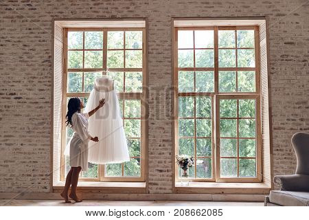 Beautiful dress. Full length of attractive young woman in silk bathrobe touching her wedding dress while standing near the window