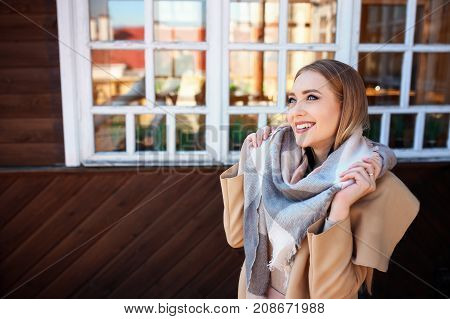 ttractive blonde woman in urban background with sun backlight. Young girl wearing winter coat and scarf standing in the street. Pretty female with straight hair hairstyle and blue eyes.