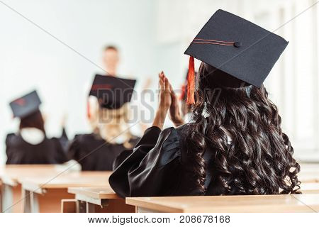 back view of student girl in graduation costume clapping hands while sitting at class