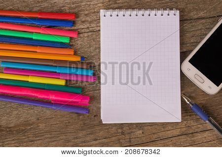 Open white notepad with colorful felt-tip pens and ball pens with smartphone on the wooden table. Space for text
