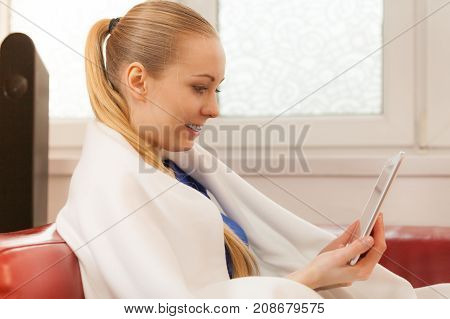 Young woman lying on sofa under warm blanket relaxing using tablet reading news and checking social media having fun during leisure time. Modern relaxation.