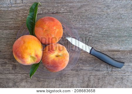 Three ripe peaches with green leaves in a plastic plate with knife on the old wooden background