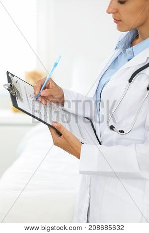 Close-up of a female doctor filling  up medical form at clipboard while standing straight in hospital.