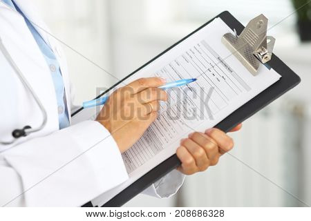 Close-up of a female doctor filling  up medical form at clipboard while standing straight in hospital.