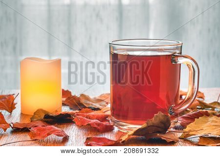 Glass cup of tea and glowing candle on wooden table with autumn leaves. Warm toning. Light tulle background