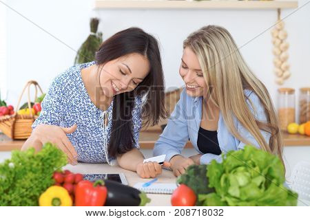 Two young happy women making online shopping for making menu by tablet computer. Friends cooking in the kitchen.