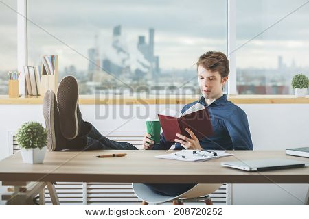 Handsome young businessman reading book and drinking coffee in modern office with blurry city view. Break concept