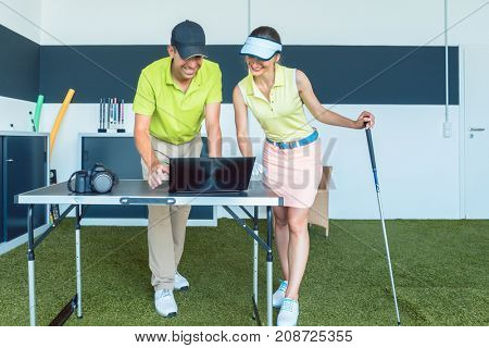 Cheerful young woman analyzing and watching on laptop her video recorded strike during a contemporary golf class with an experienced instructor