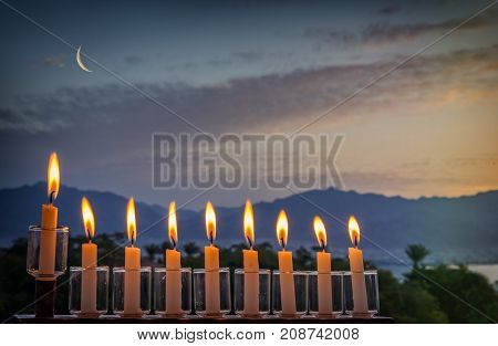 Low key image with menorah with glitter lights of burning candles and crescent moon on black sky. Image symbolizes Jewish holiday of Hanukkah