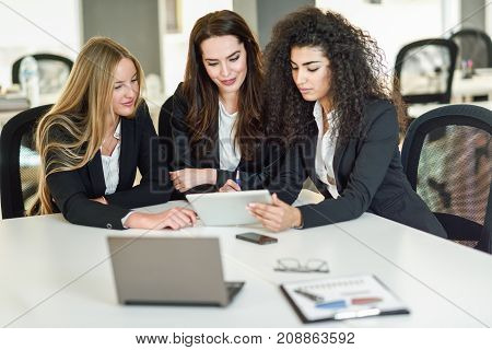 Three businesswomen working together in a modern office with white furniture. Teamwork concept. Caucasian blonde and muslim girls wearing suit. Multi-ethnic group of women