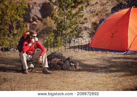Hicker with bierd and sunglasses sitting near orange tent and drink from a cup. Sunny spring daybreak in rocky mountains.