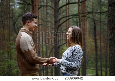 Side view of happy romantic young couple holding hands looking into each other's eyes and smiling joyfully standing among trees in autumn forest: guy declaring his love for his charming girlfriend