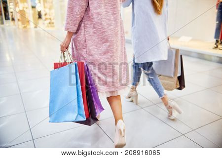 Low section portrait of two young women wearing coats and high heels walking in shopping mall holding paper bags with purchases