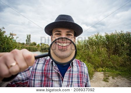 A cheerful young man with a funny face in a hat holds a magnifying glass. Let's celebrate!