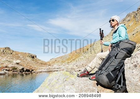 A tourist girl wearing sunglasses down jacket and hat with a backpack and mountain equipment with handles for tracking in her hands is sitting and relaxing looking at the beautiful view of the high mountain lake in the North Caucasus. Karachay-Cherkess 